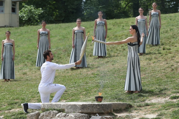 扮演最高女祭司的演員瑪麗·米娜（前右）點(diǎn)燃第一棒火炬手——希臘運(yùn)動(dòng)員、東京奧運(yùn)會(huì)男子賽艇項(xiàng)目金牌得主斯特凡諾斯·恩圖斯科斯手中的火炬。新華社記者 趙丁喆 攝